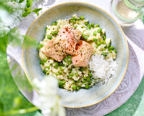 Eine Schüssel cremiges Risotto mit grünen Erbsen, belegt mit drei Stücken confiertem Lachs und garniert mit geriebenem Käse. Dieses Gericht fängt die Appetitlichkeit von Bildsprache ein und wird auf einer grün-weißen Oberfläche mit einem Glas Weißwein daneben platziert, umgeben von Grün und hellem, natürlichem Licht, perfekt für Food-Fotografie.