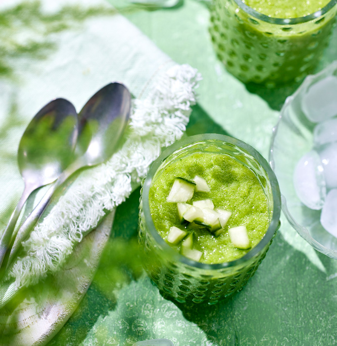 food-foto-fruehlingsgruen-gruene-gazpacho Zwei grüne Gazpacho, garniert mit kleinen Gurkenstücken, serviert in strukturierten Gläsern auf einer grünen Oberfläche. Daneben liegen Löffel auf einem weißen Tuch und überall verstreut Eiswürfel. Die Bildsprache ist hell und frisch und vermittelt ein Gefühl von Erfrischung und Kühle, perfekt für jedes Food-Fotografie-Portfolio.