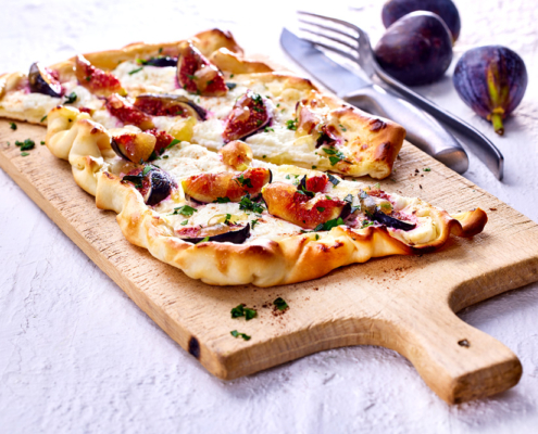 Ein frisch gebackener Flammkuchen mit geschmolzenem Ziegenkäse, Feigen und Kräutern liegt auf einem rustikalen Servierbrett aus Holz. Drei ganze Feigen und ein Besteckset sind im Hintergrund auf einer leicht strukturierten Oberfläche angeordnet und emotionalisieren die Bildsprache in diesem appetitlichen Beispiel der Food-Fotografie ein.