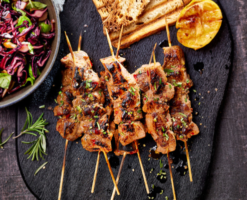 Auf einer Holzplatte liegen mehrere gegrillte Fleischspieße, garniert mit gehackten Kräutern, dazu geröstetes Brot und eine gegrillte Zitronenhälfte. Daneben steht auf einer strukturierten dunklen Oberfläche eine Schüssel mit frischem Rotkohl-Salat, begleitet von Rosmarinzweigen – eine perfekte Szene, eingefangen vom Food Fotografen Dirk Przibylla, die elegante Bildsprache zur Schau stellt.