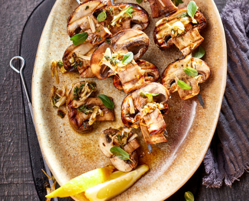 Ein Teller mit gegrillten Pilz- und Gemüsespießen, garniert mit Kräutern, wird auf einer dunklen, strukturierten Oberfläche präsentiert und verkörpert appetitliche Foodfotografie. Eine Zitronenscheibe und eine dunkle Stoffserviette liegen neben dem Teller, während ein Glas mit Kräuteröl dieses Bildsprache-Meisterwerk vervollständigt.