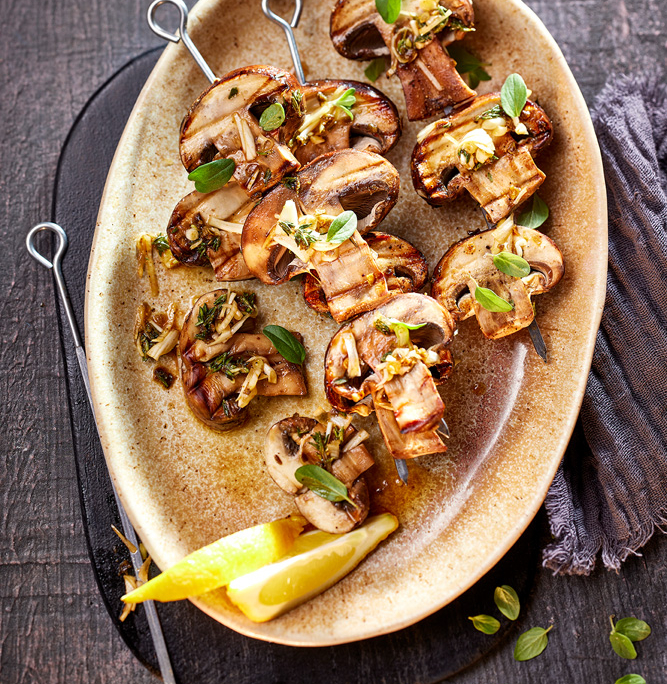 Ein Teller mit gegrillten Pilz- und Gemüsespießen, garniert mit Kräutern, wird auf einer dunklen, strukturierten Oberfläche präsentiert und verkörpert appetitliche Foodfotografie. Eine Zitronenscheibe und eine dunkle Stoffserviette liegen neben dem Teller, während ein Glas mit Kräuteröl dieses Bildsprache-Meisterwerk vervollständigt.