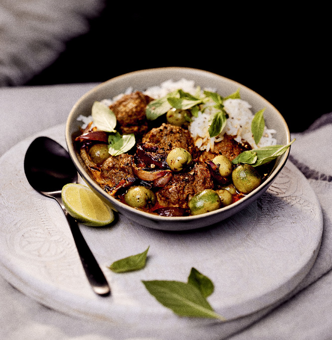 Eine Schüssel Thai-Curry auf einem weißen runden Tablett ist ein Meisterwerk der Foodfotografie. Das Curry mit Fleischbällchen, grünen Kiwibeeren und Kaffirlimettenblättern wird mit frischen Thai-Basilikum garniert und mit weißem Jasminreis serviert. Eine Limettenscheibe und ein Löffel liegen neben der Schüssel auf dem Tablett – ein wahrer Bildsprache-Moment.