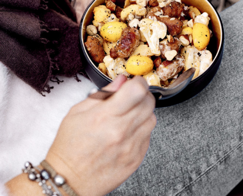 Eine Person in einem flauschigen weißen Pullover und Jeans hält eine kleine Schüssel mit buntem Gemüse. Die appetitlich gestaltete Bowl zeigt den erfahrenen Foodstylisten. Die gemütlichen Decken im Hintergrund und die entspannte Haltung der Person unterstreichen die emotionale Food-Fotografie.