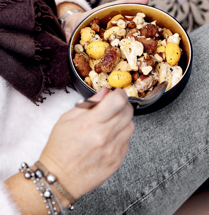 Eine Person in einem flauschigen weißen Pullover und Jeans hält eine kleine Schüssel mit buntem Gemüse. Die appetitlich gestaltete Bowl zeigt den erfahrenen Foodstylisten. Die gemütlichen Decken im Hintergrund und die entspannte Haltung der Person unterstreichen die emotionale Food-Fotografie.