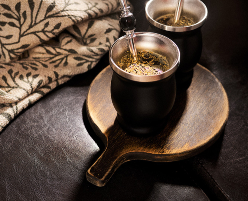 Zwei schwarze Matekürbisse mit silbernen Strohhalmen stehen auf einem hölzernen Servierbrett, das auf einem dunklen Ledersofa liegt. Neben einem Kürbis liegt eine Decke mit Blumenmuster, die eine gemütliche Atmosphäre schafft. Dampf steigt aus den Kürbissen auf, was darauf hindeutet, dass sie mit heißen Getränken gefüllt sind – ein perfekter Moment der Food-Fotografie.