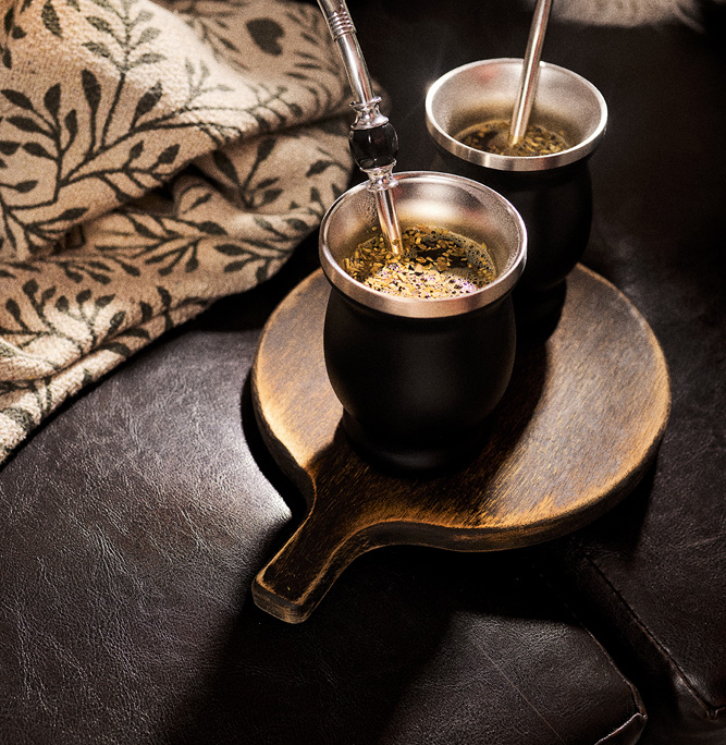 Zwei schwarze Matekürbisse mit silbernen Strohhalmen stehen auf einem hölzernen Servierbrett, das auf einem dunklen Ledersofa liegt. Neben einem Kürbis liegt eine Decke mit Blumenmuster, die eine gemütliche Atmosphäre schafft. Dampf steigt aus den Kürbissen auf, was darauf hindeutet, dass sie mit heißen Getränken gefüllt sind – ein perfekter Moment der Food-Fotografie.