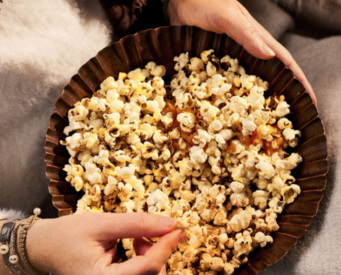 Zwei Hände halten eine Schüssel mit unterschiedlichem Popcorn. Eine Hand ruht auf dem Rand der Schüssel, während die andere hineingreift, um sich etwas Popcorn zu holen. Im Hintergrund sind eine flauschige weiße Decke und ein mit einem braunen Schal umhüllter Arm zu sehen, ein gemütlicher Moment der Foodfotografie und Bildsprache vom Food Fotografen Dirk Przibylla.