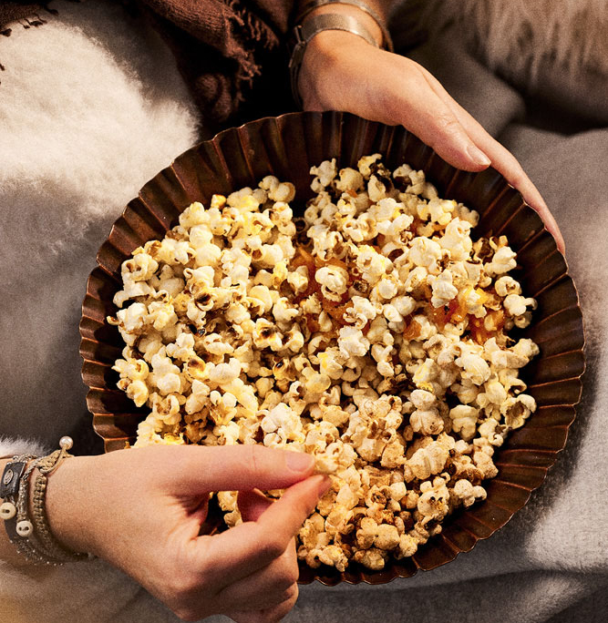 Zwei Hände halten eine Schüssel mit unterschiedlichem Popcorn. Eine Hand ruht auf dem Rand der Schüssel, während die andere hineingreift, um sich etwas Popcorn zu holen. Im Hintergrund sind eine flauschige weiße Decke und ein mit einem braunen Schal umhüllter Arm zu sehen, ein gemütlicher Moment der Foodfotografie und Bildsprache vom Food Fotografen Dirk Przibylla.