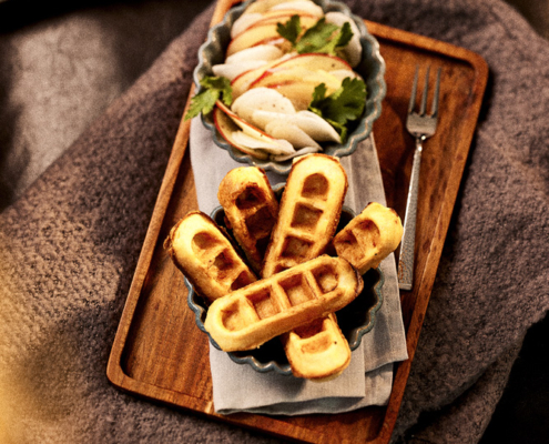 Auf einem Holztablett liegt ein Teller mit fünf fächerartig angeordneten Waffeln in Stäbchenform und eine kleine Schale mit Apfel-Rettich-Scheiben, garniert mit Petersilie. Das Tablett steht auf einer gemütlichen, dunklen Decke, neben der Schale liegt eine Gabel, die eine apptitliche Food-Fotografie abbildet und eine raffinierte Bildsprache verkörpert.