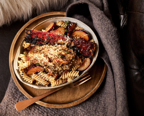 Ein Teller Spirelli mit geriebenem Käse und einem Stück gebratenem Bacon darauf, serviert auf einem Holztablett. Das Arrangement ist ein Beispiel für die exquisite Technik eines Food-Fotografen und kreative Rezeptentwicklung, arrangiert auf einer grauen Decke mit einem Fellkissen und einem dunklen Ledersofa im Hintergrund. Eine goldfarbene Gabel ist kunstvoll neben dem Teller platziert.