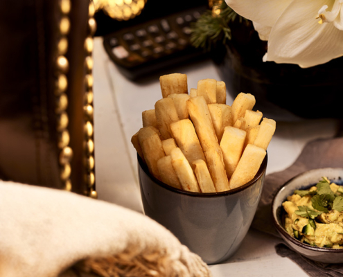 Eine kleine Schüssel mit dick geschnittenen Mainiok- Pommes frites, kunstvoll arrangiert von einem Foodstylisten, steht auf einem Beistelltisch neben einer Schüssel Avocado-Dip. Der Tisch ist mit einer gemütlichen Decke, einem großen weißen Weihnachsstern und Weihnachtsdekorationen wie goldenen Kugeln und Tanne geschmückt. Im Hintergrund ist auch eine Fernbedienung zu sehen.