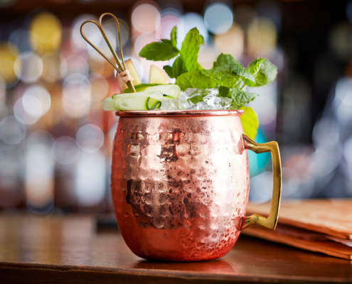 Ein kalter Moscow Mule-Cocktail, garniert mit Minzblättern, Gurkenscheiben, Ingwer und einem Rührstab, wird in einem Kupferbecher serviert, der auf einer hölzernen Bartheke steht. Der mit Food-Fotografie-Präzision aufgenommene Hintergrund ist mit Bokeh-Lichtern verwischt und schafft so eine gemütliche Atmosphäre.