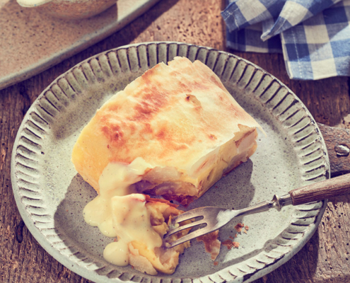 Ein quadratisches Stück Strudel mit einer flockigen, goldbraunen Kruste liegt auf einem rustikalen Teller, festgehalten in exquisiter Foodfotografie. Es wird teilweise mit einer daneben platzierten Gabel gegessen. Vanillesauce wird über den Strudel gegossen. Eine kleine Schale mit weiterer Sauce und ein blau-kariertes Tuch sind im Hintergrund und bereichern die Bildsprache.