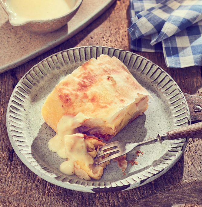 Ein quadratisches Stück Strudel mit einer flockigen, goldbraunen Kruste liegt auf einem rustikalen Teller, festgehalten in exquisiter Foodfotografie. Es wird teilweise mit einer daneben platzierten Gabel gegessen. Vanillesauce wird über den Strudel gegossen. Eine kleine Schale mit weiterer Sauce und ein blau-kariertes Tuch sind im Hintergrund und bereichern die Bildsprache.