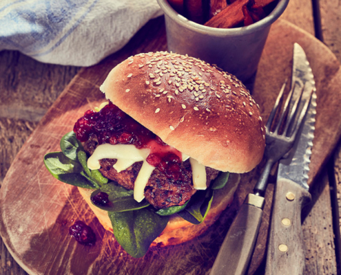 Ein Gourmet-Burger mit Spinat, Käse und Relish liegt auf einem Holzbrett neben einem Metallbecher voller Süßkartoffelpommes. Die Szene wurde von einem Foodstylisten präzise eingefangen und umfasst ein Messer, eine Gabel und ein blau-weißes Tuch auf einem rustikalen Holztisch.