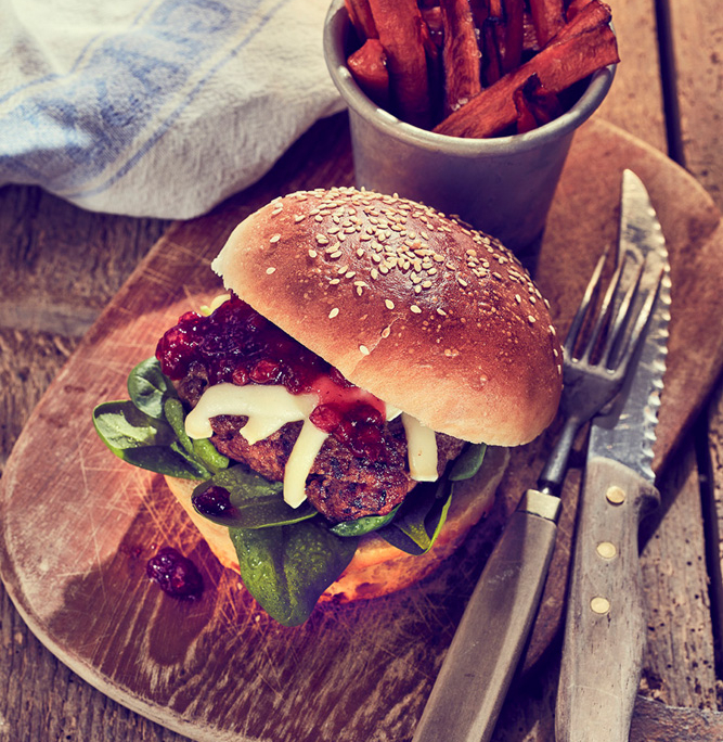 Ein Gourmet-Burger mit Spinat, Käse und Relish liegt auf einem Holzbrett neben einem Metallbecher voller Süßkartoffelpommes. Die Szene wurde von einem Foodstylisten präzise eingefangen und umfasst ein Messer, eine Gabel und ein blau-weißes Tuch auf einem rustikalen Holztisch.