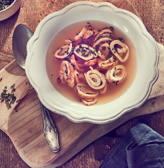 Eine rustikale Schüssel klare Suppe, garniert mit Kräutern und gefüllt mit geschnittenenPfannkuchen-Rollen, wird kunstvoll von einem Foodstylisten arrangiert. Die Schüssel steht auf einem hölzernen Schneidebrett neben einem Löffel, einer kleinen Schüssel mit Schnittlauch und einer gefalteten blauen Serviette. Dieses Food-Foto fängt die Essenz des Hausmannskosts auf einem Holztisch ein.