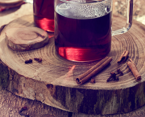 Zwei mit dunkelrotem Glühwein gefüllte Glaskrüge stehen auf einer Holzplatte. Zimtstangen und Gewürznelken sind auf der Platte verstreut und lassen die Gewürze des Getränks erahnen. Ein unscharfer Hintergrund mit einer geschliffenen Flasche und Holzstücken verstärkt das rustikale Gefühl, perfekt für Food-Fotografie.