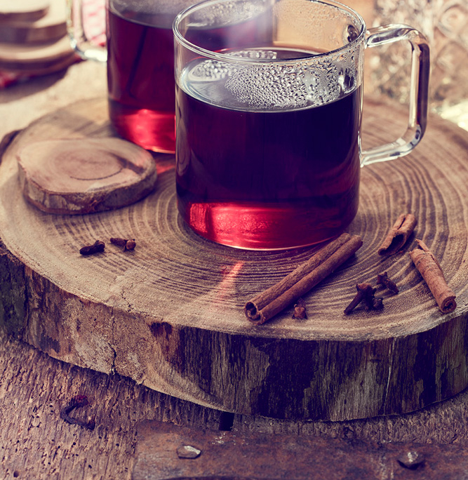 Zwei mit dunkelrotem Glühwein gefüllte Glaskrüge stehen auf einer Holzplatte. Zimtstangen und Gewürznelken sind auf der Platte verstreut und lassen die Gewürze des Getränks erahnen. Ein unscharfer Hintergrund mit einer geschliffenen Flasche und Holzstücken verstärkt das rustikale Gefühl, perfekt für Food-Fotografie.