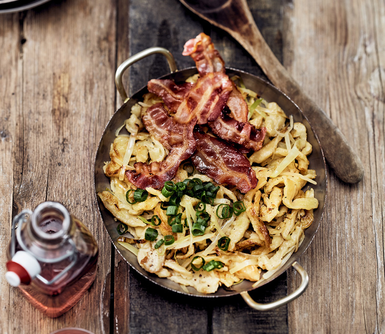 Eine Pfanne mit mehreren knusprig gebratenen Speckstreifen steht auf einer rustikalen Holzoberfläche. Ein Holzlöffel liegt neben der Pfanne. Der Speck sieht knusprig angebraten aus – eine Szene, die von einem erfahrenen Foodstylisten sorgfältig inszeniert wurde.