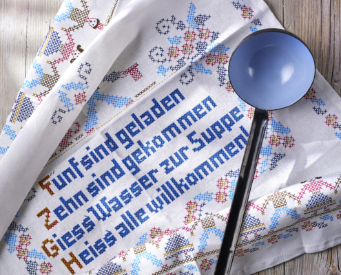 Ein dekoratives weißes Tuch mit bunter Stickerei und deutschem Text ist kunstvoll auf einer Holzoberfläche arrangiert und stellt die Essenz des Food-Designs zur Schau. Darauf ruht eine blaue Schöpfkelle mit schwarzem Griff. Der Text lautet: „Fünf sind geladen, zehn sind gekommen, gieß Wasser zur Suppe, heiß‘ alle willkommen.“.