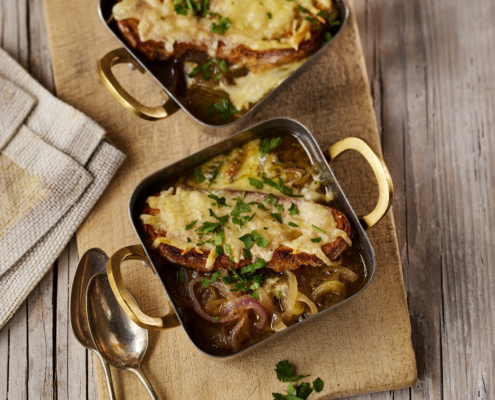 Zwei quadratische Schüsseln französischer Zwiebelsuppe, garniert mit geschmolzenem Käse und Petersilie, sind kunstvoll auf einem Holzschneidebrett arrangiert. Dieses exquisite Food-Styling wird durch eine gefaltete Serviette und zwei Löffel ergänzt, alles auf einem rustikalen Holztisch – wahrlich ein Meisterwerk der Food-Fotografie.