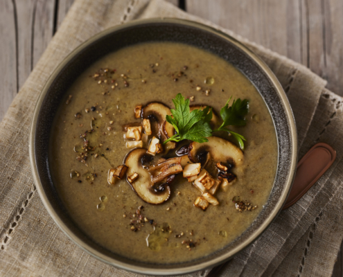 Eine Schüssel Pilzsuppe, garniert mit sautierten Pilzscheiben und Petersilie, steht elegant auf einem rustikalen Holztisch und präsentiert exquisites Food-Styling. Eine beige Stoffserviette und ein Löffel vervollständigen die Szene, während ein Zweig Petersilie dieser kunstvollen Präsentation den letzten Schliff verleiht.