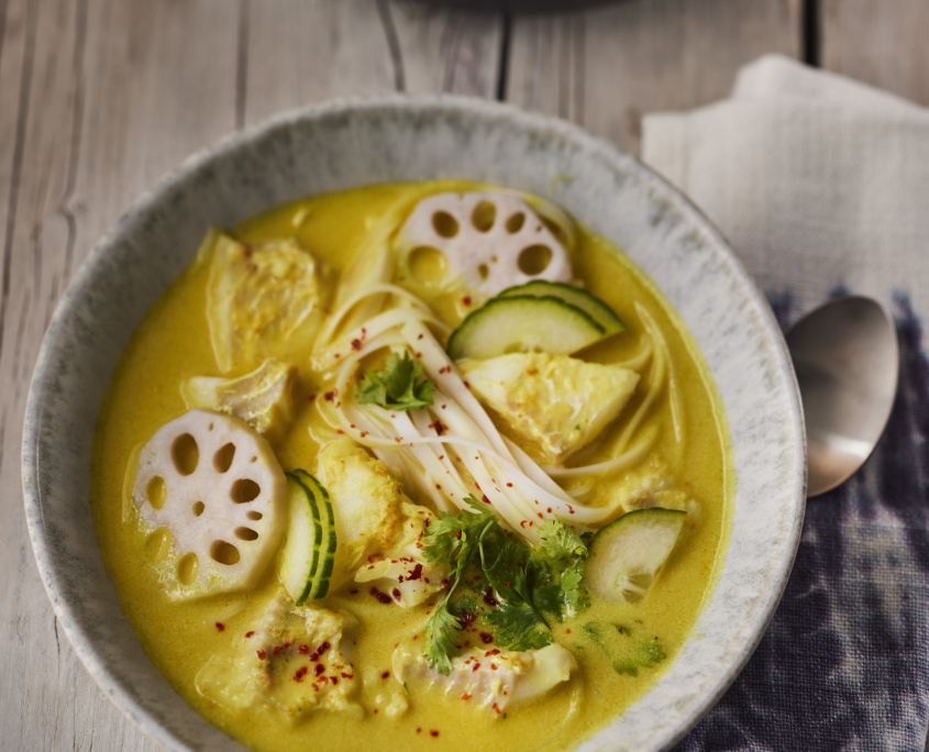 Eine Schüssel gelbe Currysuppe mit Lotuswurzelscheiben, Gurke, frischen Kräutern und Nudeln, kunstvoll auf einem Holztisch arrangiert, zeigt exquisites Fooddesign. Ein Löffel und eine Serviette liegen daneben, während ein weiteres Gericht im Hintergrund die Vision dieses Foodfotografen widerspiegelt.