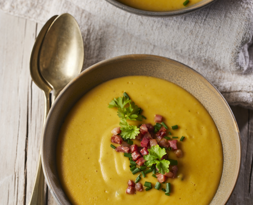 Zwei Schüsseln cremige gelbe Suppe, garniert mit gehackten Kräutern und gewürfelte Speck sind kunstvoll auf einem rustikalen Holztisch arrangiert. Dieses exquisite Food-Design zeigt einen goldenen Löffel, der neben einer Schüssel auf einer hellen Stoffserviette ruht – perfekt für die Aufnahme mit Lebensmittelfotografie.