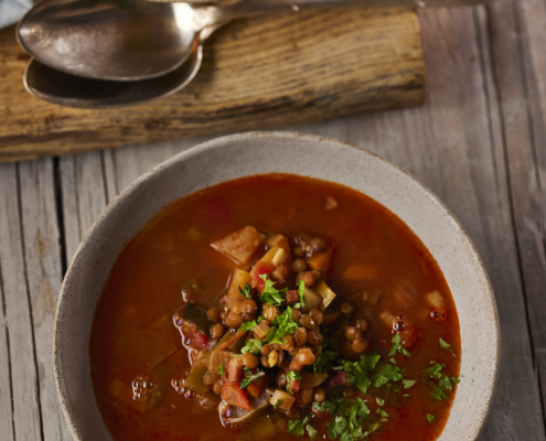 Eine Schüssel herzhafte Linsensuppe, garniert mit frischer Petersilie, steht auf einer rustikalen Holzoberfläche, mit einem Satz silberner Löffel auf einem kleinen Brett. Eine weitere Schüssel, die exquisites Foodstyling zeigt, ist teilweise im Hintergrund sichtbar und fängt die Essenz der Foodfotografie ein.