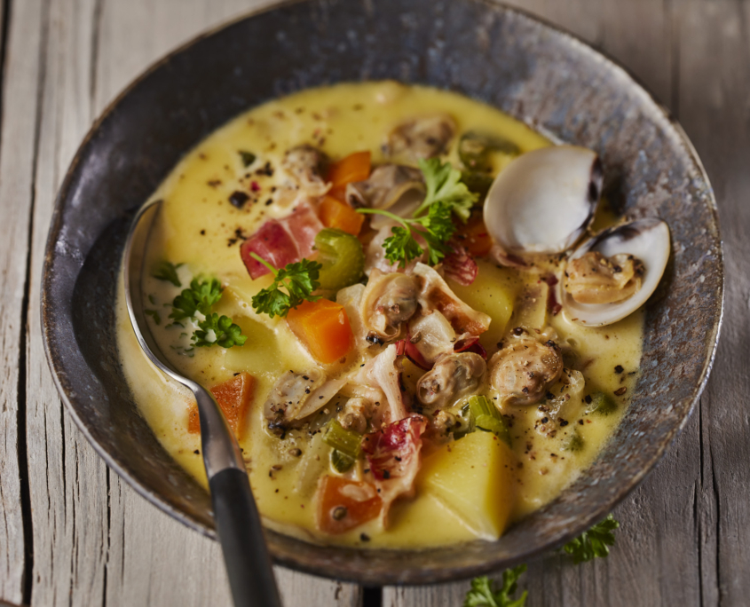 Eine rustikale Schüssel Muschelsuppe auf einem Holztisch ist ein Paradebeispiel für exquisites Foodstyling. Die cremige Suppe ist mit Muscheln, Kartoffelwürfeln und Karotten gefüllt und mit frischer Petersilie garniert. Ein Löffel liegt in der Schüssel, während ein paar Muschelschalen dieser köstlichen Komposition Charme verleihen.