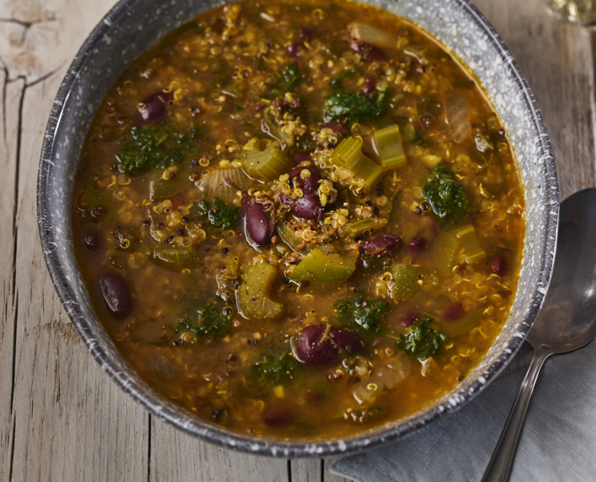 Eine Suppenschüssel mit Quinoa, Kidneybohnen, Sellerie und Blattgemüse ist kunstvoll in einer Foodfotografie-Ausstellung auf einer rustikalen Holzoberfläche festgehalten. Ein Löffel und eine gefaltete Serviette liegen neben der Schüssel, im Hintergrund steht ein Glas Wasser.