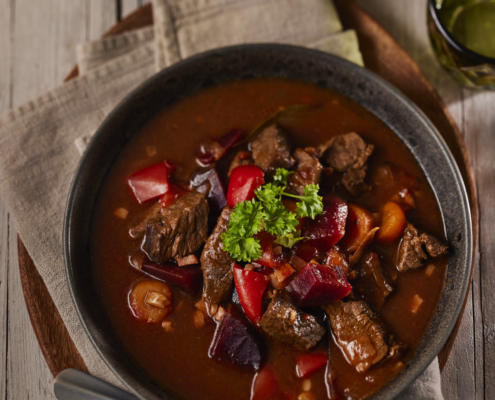 Eine Schüssel herzhafter Rindereintopf mit Rindfleischstücken, roten Paprikaschoten und Zwiebeln in kräftiger brauner Brühe, kunstvoll garniert mit frischer Petersilie. Das Gericht zeugt von tadellosem Food-Styling und steht auf einer Holzoberfläche mit einem Leinentuch und einem Löffel daneben – perfekt für jeden Food-Fotografen.