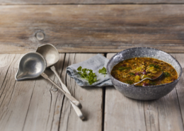 Auf einem rustikalen Tisch steht eine gesprenkelte Schüssel herzhafter Gemüsesuppe, garniert mit frischen Kräutern. Zwei antike silberne Schöpfkellen und eine graue Serviette mit Petersilie liegen neben der Schüssel auf dem hölzernen Hintergrund und sind eine Hommage an außergewöhnliches Food-Design. Sie verkörpern elegante Lebensmittelfotografie.