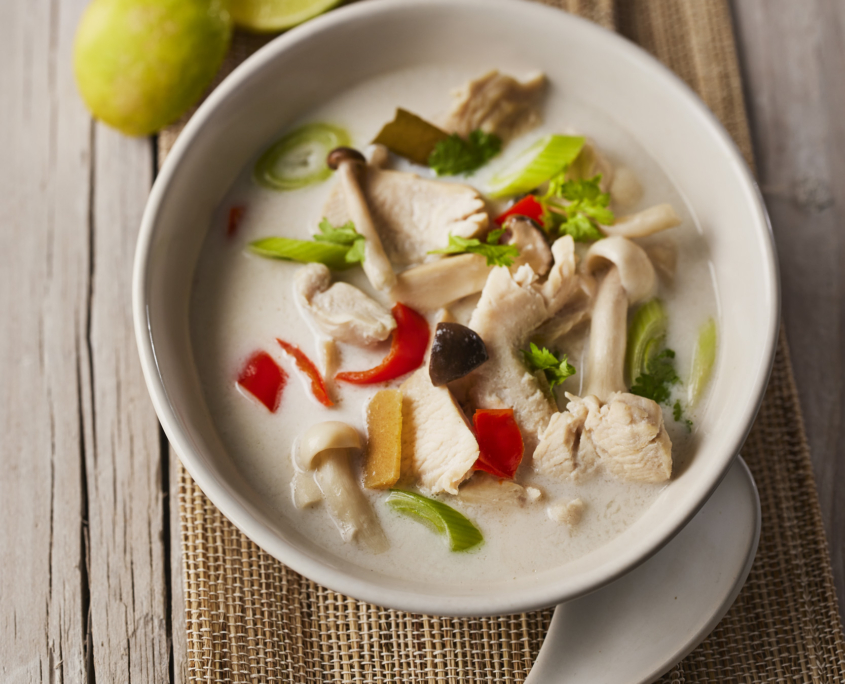 Eine Schüssel cremige Suppe mit geschnittenen Champignons, Hühnchen, roten Chilis und Frühlingszwiebeln, garniert mit Koriander – perfekt für Food-Design-Enthusiasten. Ein weißer Keramiklöffel liegt neben der Schüssel, während im Hintergrund auf einer rustikalen Holzoberfläche eine halbierte Limette liegt. Ideal für alle, die sich für Food-Fotografie oder Lebensmittelfotografie interessieren.