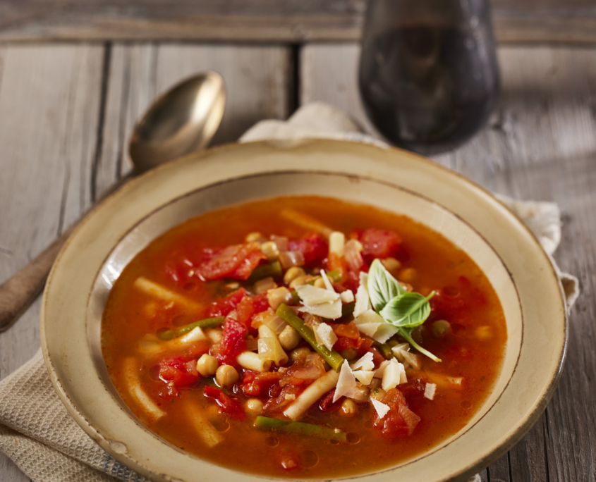 Eine rustikale Schüssel Gemüsesuppe mit Tomaten, Kichererbsen und Kräutern, garniert mit frischem Basilikum und geriebenem Käse. Ein Löffel und ein Glas auf einem Holztisch fangen die Essenz der Lebensmittelfotografie ein und unterstreichen die gemütliche, rustikale Atmosphäre.