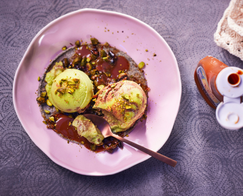 Ein rosa Teller mit drei Kugeln grünem Eis, mit Avocadogeschmack, beträufelt mit Schokoladensauce und garniert mit zerstoßenen Nüssen und Trockenfrüchten. Auf dem Teller liegt ein Löffel. Auf der rechten Seite des Bildes ist eine Flasche Karamellsauce zu sehen, die auf einer strukturierten lila Oberfläche steht und von erfahrener Food-Fotografie stammt.