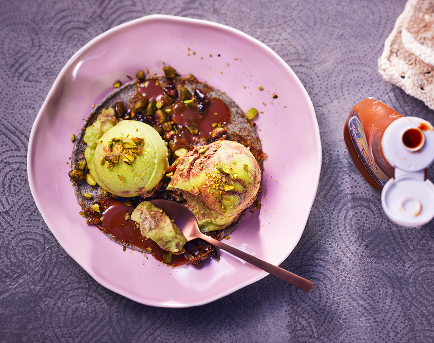 Ein rosa Teller mit drei Kugeln grünem Eis, mit Avocadogeschmack, beträufelt mit Schokoladensauce und garniert mit zerstoßenen Nüssen und Trockenfrüchten. Auf dem Teller liegt ein Löffel. Auf der rechten Seite des Bildes ist eine Flasche Karamellsauce zu sehen, die auf einer strukturierten lila Oberfläche steht und von erfahrener Food-Fotografie stammt.