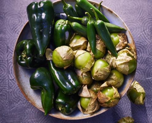 Eine Schüssel gefüllt mit verschiedenen grünen Paprikasorten, darunter dunkelgrüne Poblano-Paprika, etwas heller grüne Jalapeño-Paprika und kleine grüne Tomatillos in papierartigen Schalen. Die kunstvolle Bildsprache fängt die leuchtenden Farben perfekt auf der strukturierten lila Oberfläche ein und zeigt gekonnte Food-Fotografie.