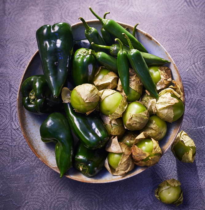 Eine Schüssel gefüllt mit verschiedenen grünen Paprikasorten, darunter dunkelgrüne Poblano-Paprika, etwas heller grüne Jalapeño-Paprika und kleine grüne Tomatillos in papierartigen Schalen. Die kunstvolle Bildsprache fängt die leuchtenden Farben perfekt auf der strukturierten lila Oberfläche ein und zeigt gekonnte Food-Fotografie.