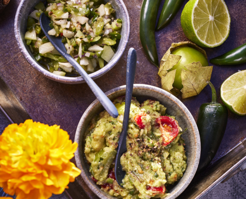 Eine lebendige Essensszene, eingefangen in atemberaubender Foodfotografie: zwei Schalen mit Dips – eine gefüllt mit stückiger Guacamole, garniert mit Kirschtomaten und Gewürzen, die andere mit grüner Salsa. Frische grüne Chilis, halbierte Limetten, Tomatillos und leuchtend gelbe Ringelblumen umgeben die Schalen auf einem rustikalen Tablett.