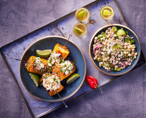 Auf einem Tablett steht ein Teller mit gegrilltem Maiskolben, garniert mit Käse und Kräutern und Limettenspalten. Daneben steht eine Schüssel mit gemischtem Bohnen-Gemüse-Salat mit Fetakäse. Drei kleine Gläser mit Teqila und Limette und zwei kleine Chilischoten stehen daneben, eingefangen mit exquisiter Food-Fotografie.