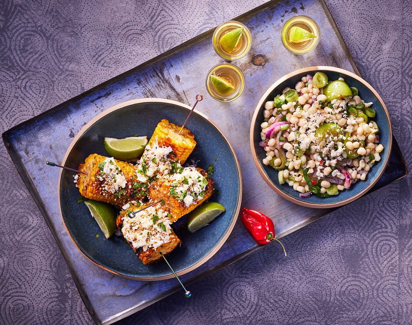 Auf einem Tablett steht ein Teller mit gegrilltem Maiskolben, garniert mit Käse und Kräutern und Limettenspalten. Daneben steht eine Schüssel mit gemischtem Bohnen-Gemüse-Salat mit Fetakäse. Drei kleine Gläser mit Teqila und Limette und zwei kleine Chilischoten stehen daneben, eingefangen mit exquisiter Food-Fotografie.