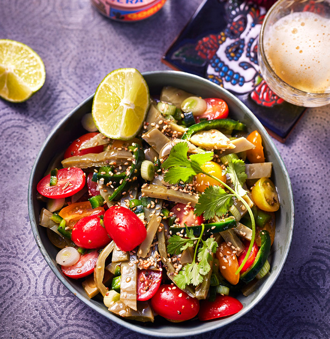 Ein farbenfroher Salat in einer Schüssel mit Kirschtomaten, Koriander, Sesamkörnern, Nopales und Limettenspalten an der Seite, begleitet von einer Dose mexikanischem Bier, einem Glas und Dekorationsartikeln, darunter Blumen und einem Untersetzer mit Tag-der-Toten-Motiv – wunderschön eingefangen von einem erfahrenen Foodstylisten.