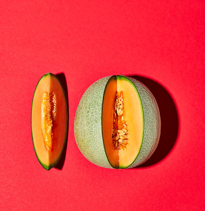 Eine angeschnittene Cantaloupe-Melone, bei der eine Hälfte die Kerne zeigt und die andere Hälfte in eine Scheibe geschnitten ist, auf einem leuchtend roten Hintergrund. Die grüne Außenschale kontrastiert mit dem orangefarbenem Fruchtfleisch und zeigt eine exquisite Bildsprache.