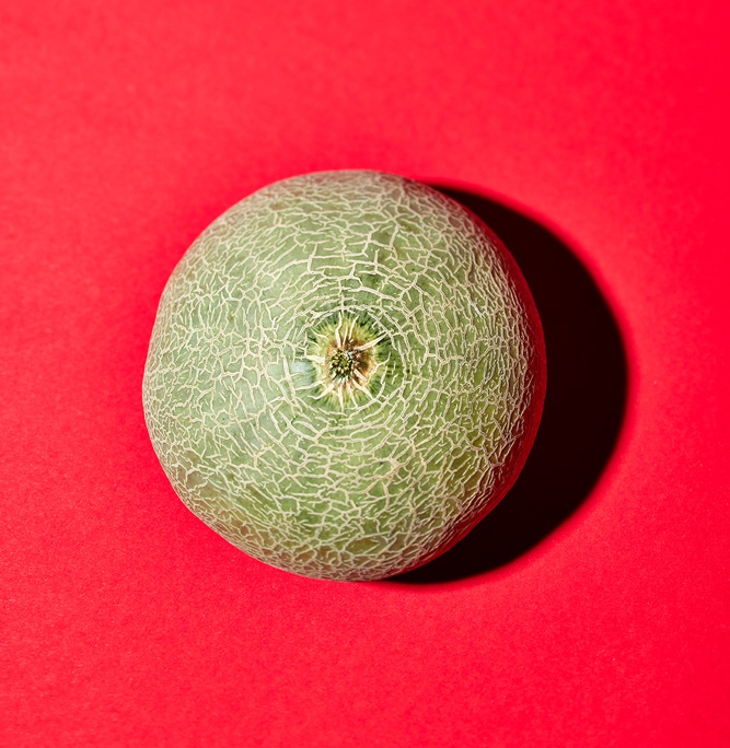 In diesem Beispiel für Food-Fotografie steht eine ganze, strukturierte grüne Cantaloupe-Melone vor einem knallig roten Hintergrund. Die Melone wirft einen Schatten auf eine Seite, wodurch ein auffälliger Kontrast zum hellen Hintergrund entsteht und die Bildsprache emotionalisiert wird.