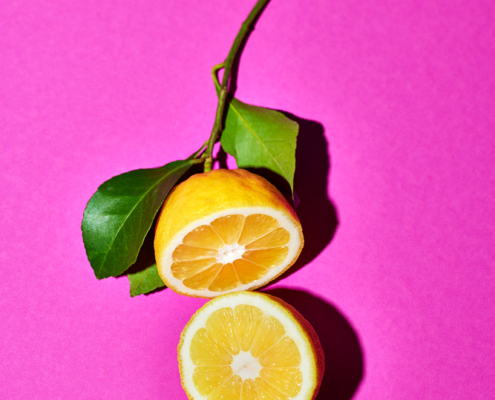 Eine halbierte Amalfizitrone liegt auf einer pinken Oberfläche, wobei beide Hälften flach liegen und ihr saftiges Inneres zeigen. Eine Hälfte ist noch an einem kleinen Stängelstück mit zwei grünen Blättern. Die leuchtenden Farben bilden einen scharfen Kontrast zum pinken Hintergrund und zeigen die Kunst eines sorgfältigen Foodfotografen.