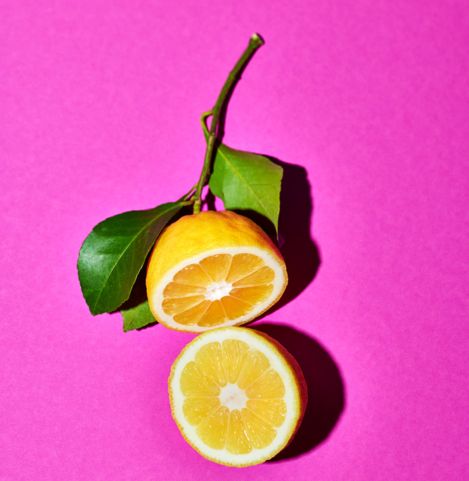 Eine halbierte Amalfizitrone liegt auf einer pinken Oberfläche, wobei beide Hälften flach liegen und ihr saftiges Inneres zeigen. Eine Hälfte ist noch an einem kleinen Stängelstück mit zwei grünen Blättern. Die leuchtenden Farben bilden einen scharfen Kontrast zum pinken Hintergrund und zeigen die Kunst eines sorgfältigen Foodfotografen.