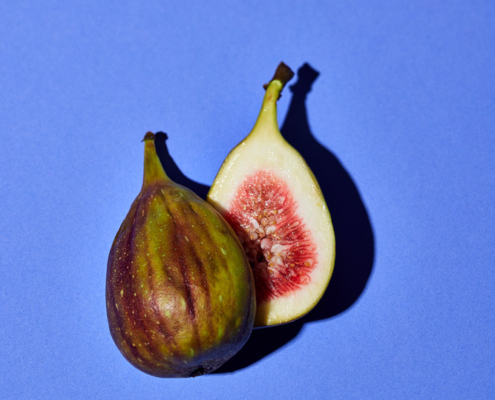 Eine halbierte Feige wird vor einem violetten Hintergrund präsentiert, fachmännisch arrangiert von einem Foodstylisten. Die eine Hälfte der Feige hat eine grün-violett gestreifte Schale, während die andere Hälfte ihr rosa, mit Samen gefülltes Inneres offenbart. Die kontrastierenden Farben lassen die Früchte in dieser atemberaubenden Lebensmittelfotografie hervorstechen.